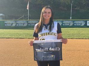 a portrait photograph of Jamie Kelly, standing on the wvu potomac state college softball field and holding the catamounts "Climb Higher" banner on a bright day